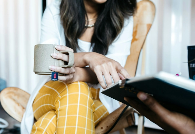 woman in white long sleeve shirt and yellow and white plaid pants sitting on chair drinking tea